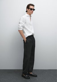 Un uomo sta in piedi con sicurezza contro un muro grigio chiaro, indossando una camicia bianca, pantaloni neri e occhiali da sole, con le mani nelle tasche.