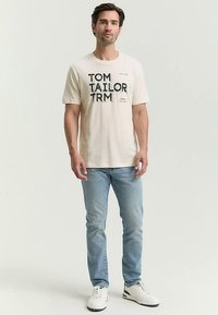 Kremfarvet t-shirt med sort grafisk tekst "TOM TAILOR" foran. Slanke jeans og hvide sneakers fuldender det afslappede look.