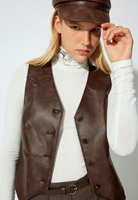 Bruine nep leren gilet met een slim fit ontwerp, puntige zoom en vijf knopen. Draagt over een witte coltrui en grijze broeken.
