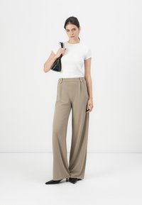 Woman standing in beige wide-leg trousers, white short-sleeve top, black pointed shoes, carrying black handbag on shoulder against plain background.
