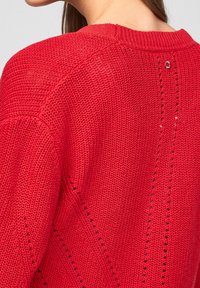 Maglione rosso lavorato a maglia con un motivo testurizzato e piccoli ritagli, colletto a coste e un accentuato rotondo sul retro.