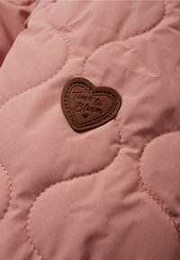 Hnědá náplast ve tvaru srdce s nápisem "Čas rozkvést" vyraženým na růžové prošívané tkanině s vlnkovým stehem.