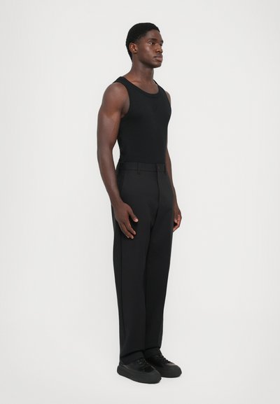 Schwarzes Tanktop, maßgeschneiderte schwarze Hose, glattes Material, schlankes Design und schwarze Schuhe, präsentiert an einem männlichen Modell vor einem weißen Hintergrund.