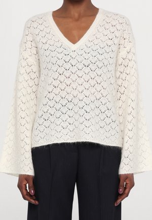 Pull blanc en tricot avec col en V, motif en losanges et manches larges, porté avec un pantalon noir sur une personne à peau foncée, montré des épaules aux hanches.
