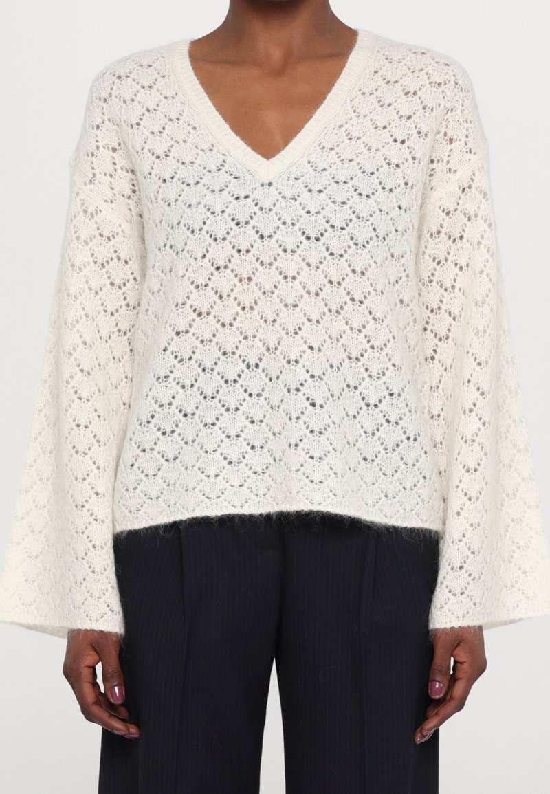 Pull blanc en tricot avec col en V, motif en losanges et manches larges, porté avec un pantalon noir sur une personne à peau foncée, montré des épaules aux hanches.