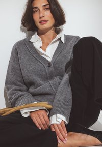 Grau gestrickte Strickjacke mit V-Ausschnitt, ausgestattet mit einem einzelnen Knopf. Darunter ein weißes Hemd mit Kragen, kombiniert mit schwarzen Hosen. Minimalistische Umgebung.