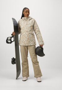 Skijacke mit beigem Blumenmuster, beigefarbene Hose und pinke Stiefel. Hält ein schwarzes Snowboard und einen schwarzen Helm. Glatte Textur, Kapuzendesign.