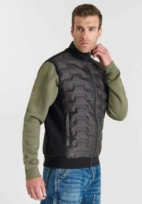 Gilet imbottito con corpo nero, maniche verde oliva con motivo testurizzato e colletto alto. Chiusura con zip e tasche laterali.