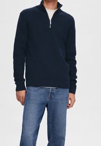 Laivastonsininen vetoketjullinen neulepusero, jossa on teksturoiduista neulekankaasta valmistettu, resorit hihansuissa ja helmassa, yhdistetty vaaleansinisiin farkkuihin.