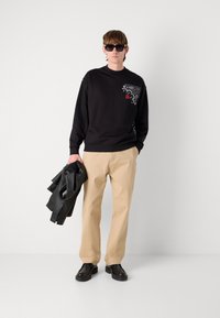 Sudadera negra con estampado floral blanco y logotipo rojo, combinada con pantalones beiges y zapatos de cuero negros. El modelo sostiene una chaqueta de cuero negra.