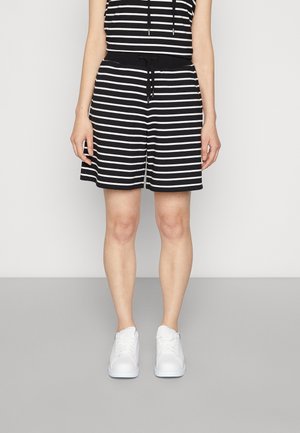 Personne portant un short et un haut rayés horizontaux noirs et blancs, assortis à des baskets blanches, debout devant un fond uni.
