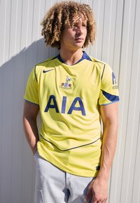 Gul sports jersey med blå accenter og "AIA" logoen præsenteret iøjnefaldende. Har rund hals og korte ærmer. Athletic-fit, glat tekstur.