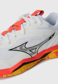 Baskets de sport blanches avec tige en maille, logo noir, accents orange et semelle jaune. Comprend des motifs texturés et un système de laçage robuste.
