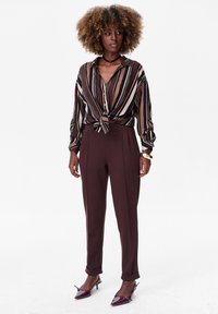 Chemise à rayures avec un devant noué, associée à un pantalon tailleur marron et des escarpins bordeaux à bouts pointus, mettant en valeur des textures lisses.