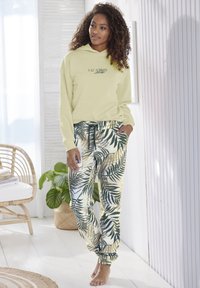 Gele hoodie met de tekst "VACATION", gecombineerd met joggers met een groen en gouden bladpatroon. Materiaal lijkt zacht; relaxte pasvorm, met een elastische tailleband.