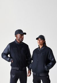 Duas pessoas vestem jaquetas varsity pretas do Chicago Bulls e bonés combinando, pousando contra um fundo claro e simples.