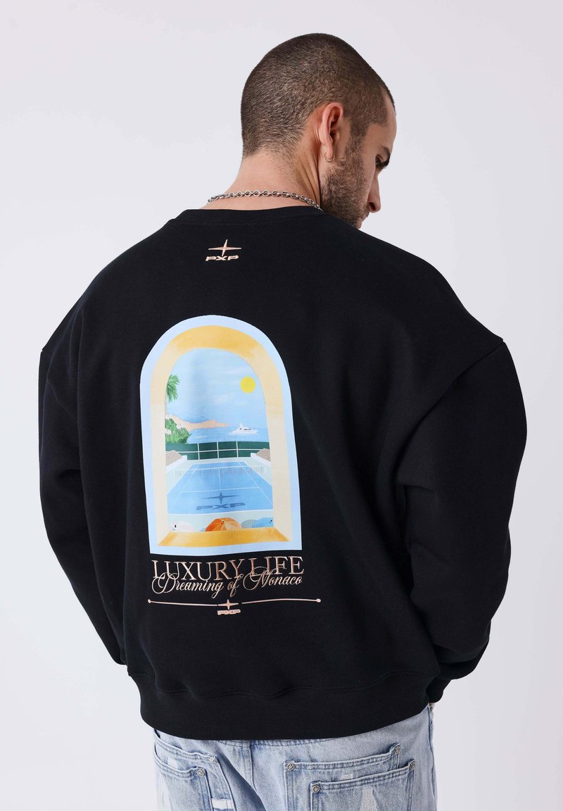 Homme en sweat noir avec un graphique de fenêtre colorée représentant une piscine, la mer et le soleil, avec le texte "Luxury Life Dreaming of Monaco."
