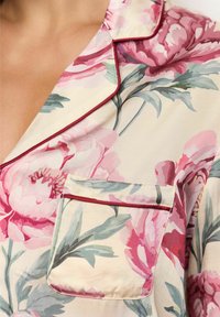 Top de pijama floral din țesătură crem, cu un model mare de bujori roz. Dispune de un buzunar pe piept cu bordură roșie și detaliu cu țesătură pe guler.