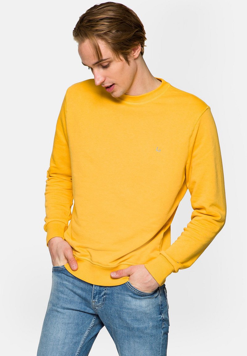 Żółty bawełniany sweatshirt z okrągłym dekoltem i ściągaczami przy mankietach, połączony z niebieskimi dżinsami. Ma małe logo na klatce piersiowej.