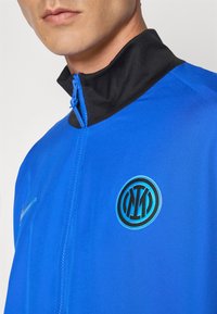 Blå atletisk jakke med høj sort krave, der har et rundt logo og en frontlynlås. Glat stof med et sporty design.