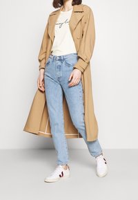Un trench-coat beige, un t-shirt crème avec un texte noir, un jean bleu clair et des baskets blanches avec des accents bordeaux. Tissu doux et coupe décontractée.