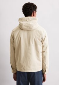 Beżowa kurtka z kapturem o gładkiej fakturze, z detalem w postaci szwu z tyłu, regulowanymi mankietami oraz luźnym krojem.