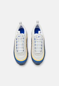 Tenisky Nike s vrstveným designem, s bílou síťovinou, modrými a žlutými akcenty a texturovanou gumovou podrážkou. Horní pohled ukazuje tkaničky a logo.