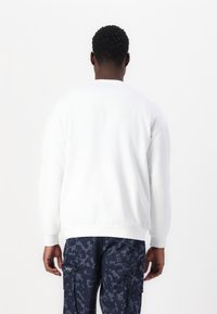 Vit crewneck sweatshirt med långa ärmar och ribbade manschetter, med en strukturerad tyg och elastisk nederkant. Buren tillsammans med mönstrade marinblå cargobyxor.