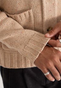 Pleteni cardigan v bež barvi z večbarvnimi pikami, z ribastimi manšetami in sprednjim žepom. Opazna podrobnost iz lesenih gumbov na zapenjanju.