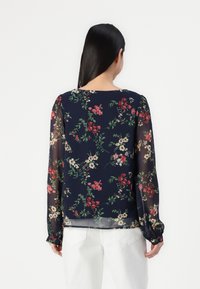 Chemisier fleuri sur fond marine, manches longues transparentes, encolure ronde et motifs de fleurs colorées en rouge, blanc et vert.
