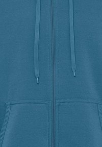Téglalap alakú, zipzáros fleece kapucnis pulóver türkiz színben, sima textúrával, állítható húzózsinórokkal és kenguruzsebekkel. Teljes hosszúságú elülső zipzárral rendelkezik.