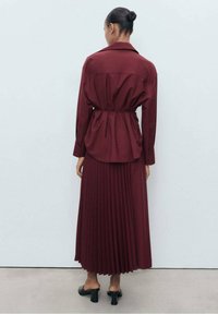 Maglietta bordeaux con gonna midi plissettata, caratterizzata da una vita stretta e maniche lunghe. Tessuto lavorato, design elegante e scarpe con tacco.