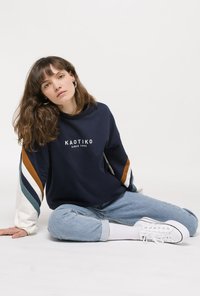 Mørkeblå sweatshirt med hvide, teal- og brune stribede ærmer og "KAOTIKO" logo. Parret med lyseblå jeans og hvide sneakers.