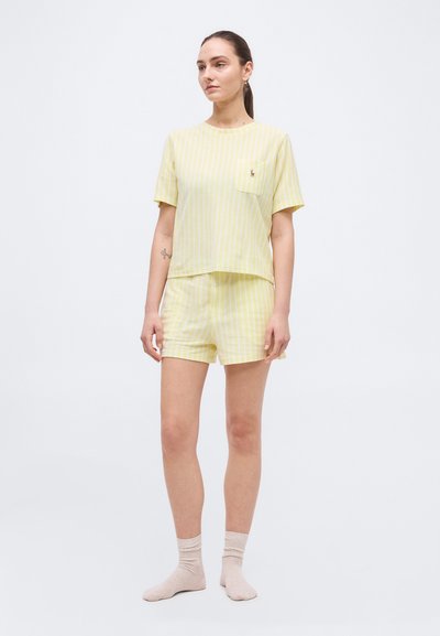 Νεαρή γυναίκα που στέκεται, φοράει κίτρινη και λευκή ριγέ κοντομάνικη μπλούζα και ταιριαστό σορτς, με ανοιχτόχρωμες κάλτσες, μπροστά σε απλό φόντο.