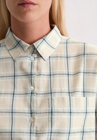 Kobieta w beżowej i jasnoniebieskiej koszuli w kratkę z guzikami, z kołnierzykiem i kieszonką na piersi, pokazana z widokiem od podbródka do połowy torsu.