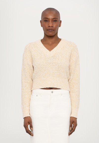 Beige verkürzter Pullover mit strukturiertem Strick, V-Ausschnitt und gerippten Bündchen. Kombiniert mit einem weißen Denimrock, der mit einem Knopfverschluss und vorderen Taschen versehen ist.