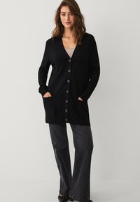 Zwarte lange cardigan met een V-hals, knoopsluiting aan de voorkant en twee voorkruiden. Zachte textuur, middellange uitstraling, gecombineerd met grijze wijde broek.