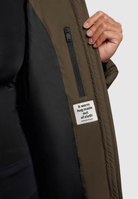 Veste olive à l'intérieur comportant une doublure noire, une poche zippée et une étiquette indiquant "Un câlin chaleureux en tissu." Texture lisse.
