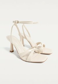 Sandales à talons hauts beiges avec de fines lanières, bouts carrés et finition brillante. Elles possèdent un talon carré et une lanière de cheville réglable.
