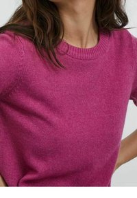 Femme portant un pull en tricot rose vif à manches courtes avec un col rond, visible des épaules à la taille.