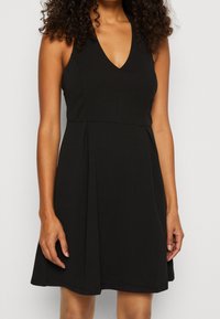 Czarna sukienka bez rękawów z głębokim dekoltem w kształcie litery V, dopasowaną górą i zwiewną spódnicą, wykonana z gładkiego materiału z delikatnymi szwami i luźnym krojem.