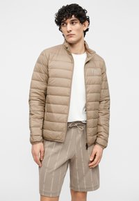 Barna steppelt pufidzseki álló gallérral és cipzáras elülső résszel. Bézs színű, mintás rövidnadrággal párosítva, amely nyomtatott szöveget és húzózsinóros megoldást tartalmaz.