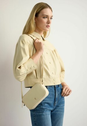 Blondynka w beżowej, zapinanej na guziki kurtce i niebieskich dżinsach, trzymająca małą beżową torebkę na ramię.