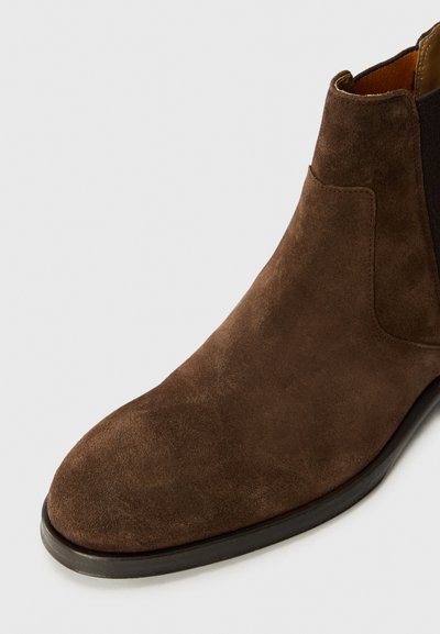Bota de tobillo de ante marrón con parte superior suave, paneles laterales elásticos y puntera redondeada. Suela de goma negra que proporciona durabilidad y agarre.