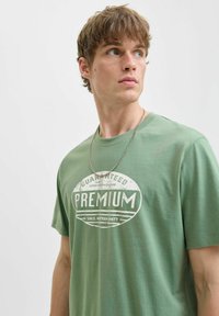 Žalias medvilninis marškinėliai trumpomis rankovėmis ir apvalia iškirpte. Pasižymi baltu "PREMIUM" grafinio spaudinys ir sidabriniu grandininiu kaklo papuošalu.