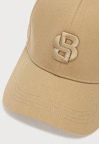 Beige baseball-lippis, joka on valmistettu teksturoidusta kankaasta ja jossa on kohotettu, kirjailtu logo samansävyisenä. Kaareva lippa, jossa näkyvät ompelusaumat.