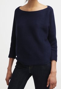 Námorně modrý pletený svetr s lodičkovým výstřihem, tříčtvrtečními rukávy a vodorovným žebrovaným vzorem, doplněný volným střihem.