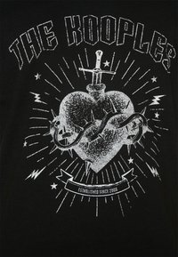 Camiseta negra con un gráfico de un corazón con una espada y alambre de espino, rodeado de relámpagos y estrellas; texto "THE KOOPLES" en la parte superior.