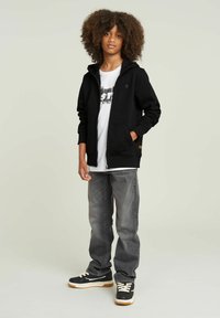Sudadera negra con cremallera y bolsillos sobre una camiseta gráfica blanca, combinada con jeans grises y zapatillas negras con suela crema.