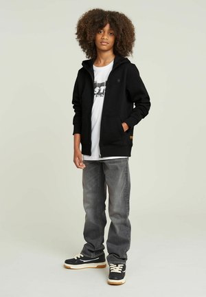 Felpa con zip nera con tasche sopra una t-shirt bianca con grafica, abbinata a jeans grigi e sneaker nere con suola crema.